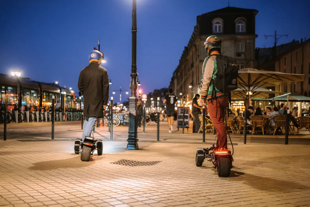 Two riders seen from behind in the evening, in a lit street lined with café terraces