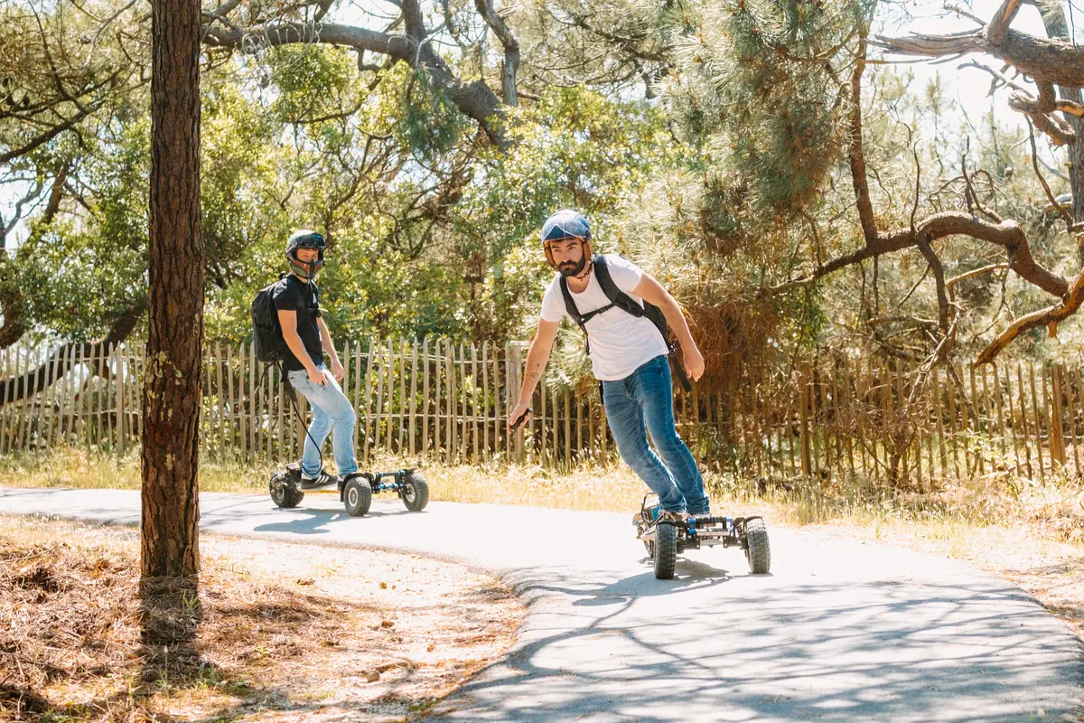 Two helmeted riders on a narrow road through the forest, alongside a fence