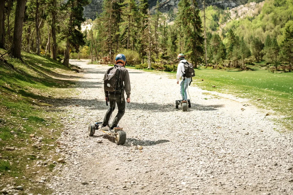 Two riders seen from behind on a gravel mountain track, among the fir trees